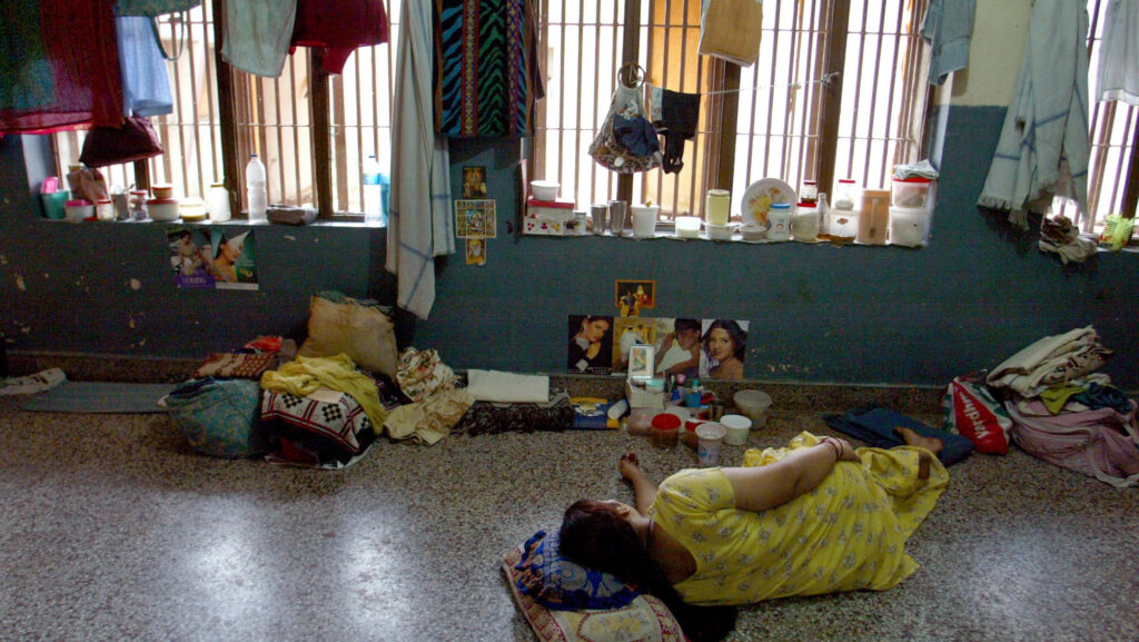 Woman in a yellow dress lying on the floor of a crowded, sparse prison cell with laundry hanging on barred windows and belongings scattered around
