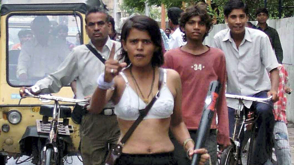 A woman walks defiantly through a busy street in India, raising a finger and carrying a stick, surrounded by onlookers and cyclists