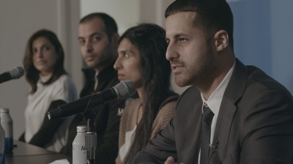 Quatro participantes sentados à mesa com microfones durante um debate, com um homem de terno falando em primeiro plano. Quatro participantes sentados à mesa com microfones durante um debate, com um homem de terno falando em primeiro plano.