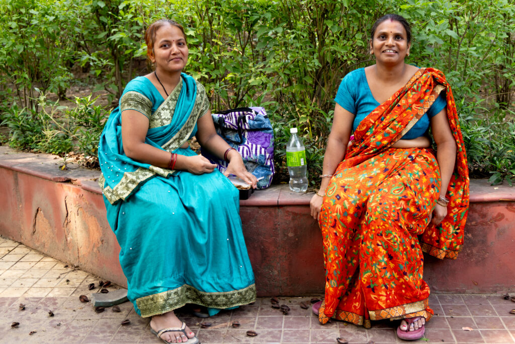 Two women in colorful saris sitting together on an outdoor ledge surrounded by greenery