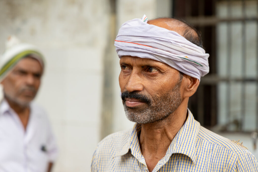 Man wearing a white cloth head wrap with orange and green stripes, looking to the side, with another man blurred in the background