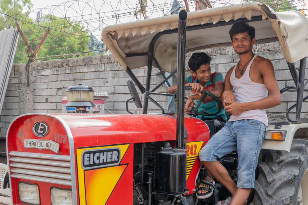 Two young men sitting on a red Eicher tractor, smiling, with a concrete wall and barbed wire fence in the background