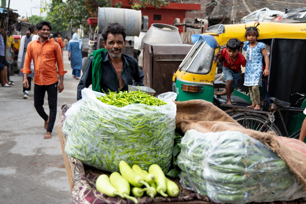 A street vendor stands behind a cart loaded with large bags of green chilies and other vegetables on a busy urban street in India