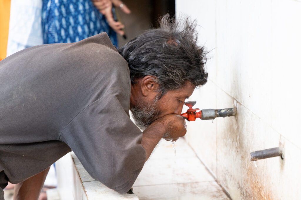 A man bends down to drink directly from an outdoor wall-mounted faucet, water dripping from his mouth
