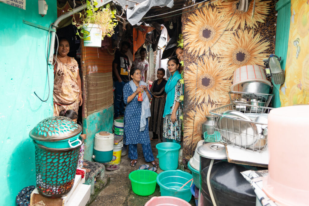 A group of women gathered in a narrow alleyway of an urban informal settlement in India, surrounded by household items and colorful walls