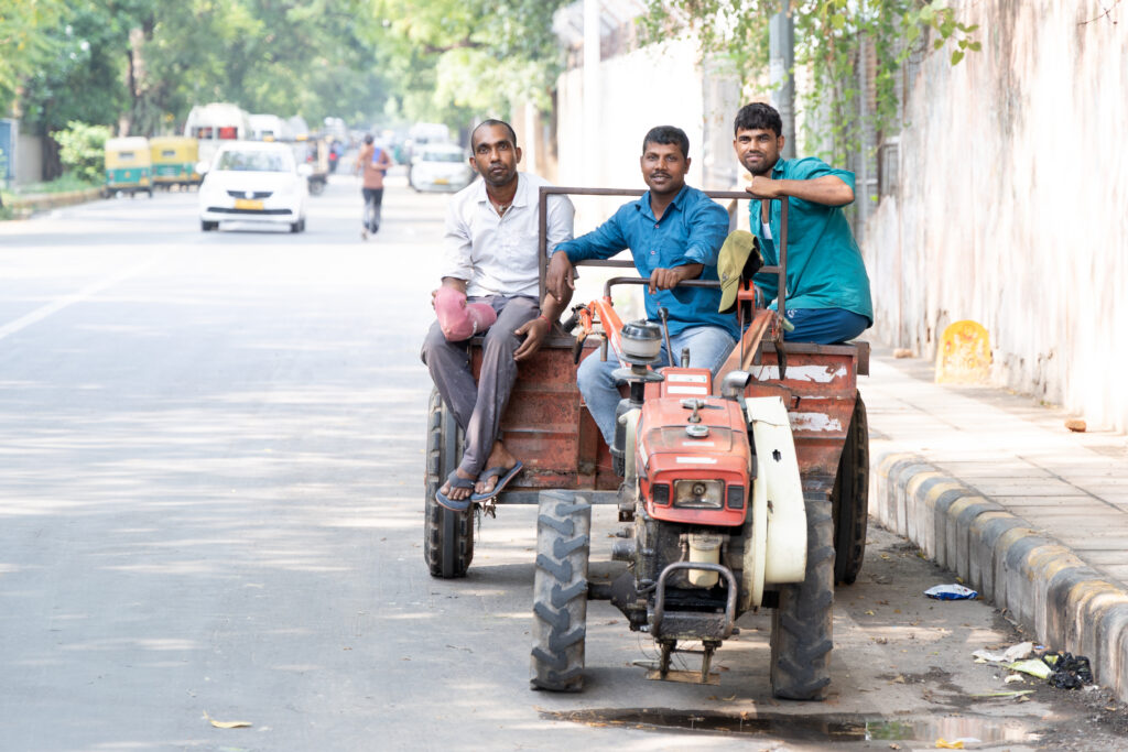 Three men sitting on a small red tractor parked along a busy urban street in India