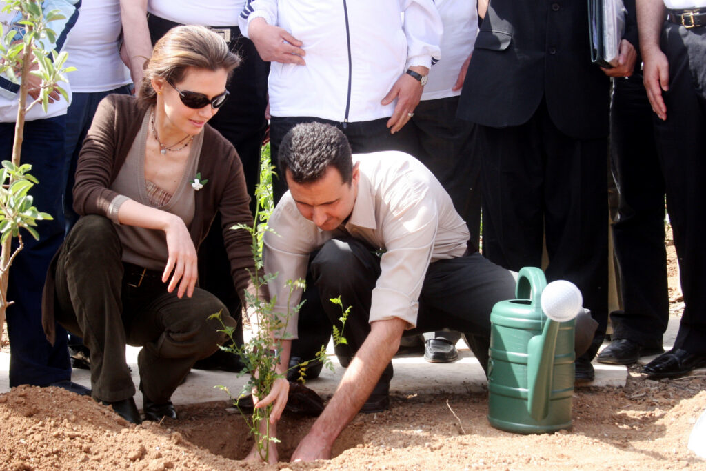 A man and woman crouch together to plant a small tree in soil, with a green watering can nearby and onlookers standing behind them