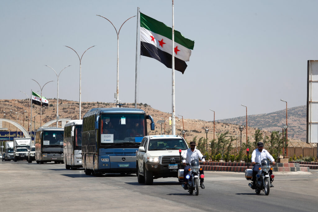 A convoy of IOM and UNHCR buses escorted by motorcycle police passes under Syrian opposition flags along a desert highway