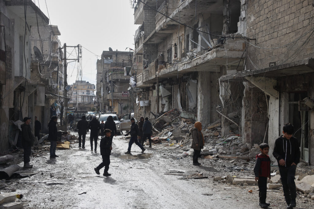 People walk through a war-damaged street lined with destroyed and partially collapsed buildings, rubble scattered across the road