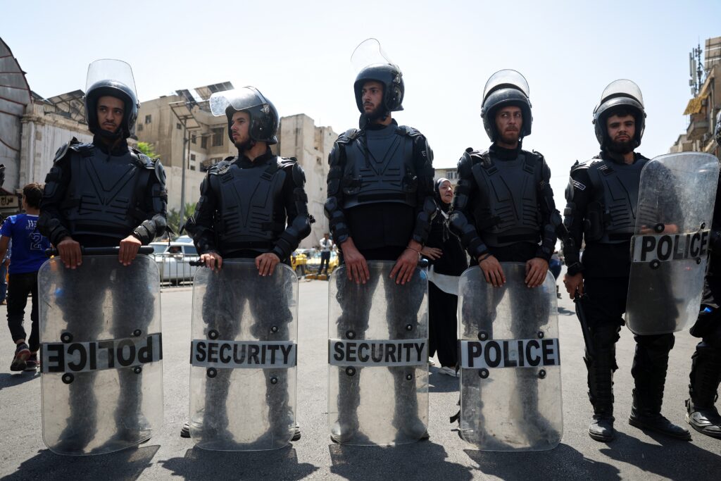 Riot police and security officers in full gear and helmets hold shields while forming a line on a street