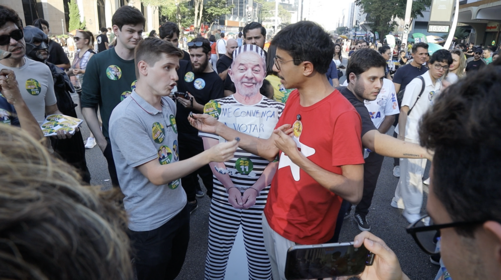 Two men debate at a Brazilian political rally near a cardboard cutout of Lula in a prison uniform reading