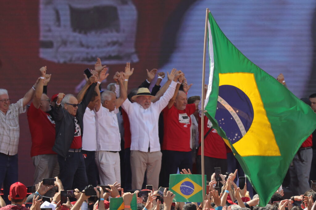 A group of politicians raise joined hands on stage at a rally, with Brazilian flags waving in the foreground crowd