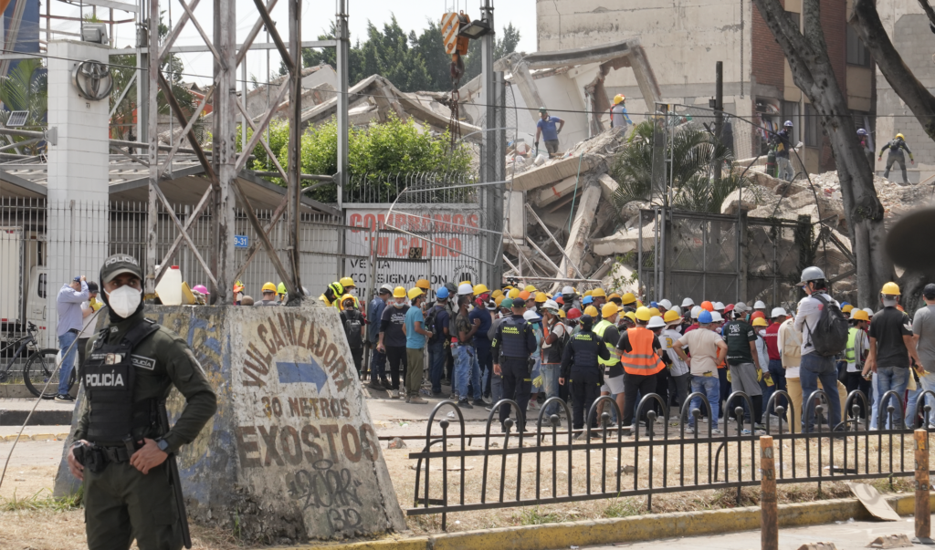 Colombian police officer and rescue workers in hard hats gather near a collapsed building, with debris visible in the background