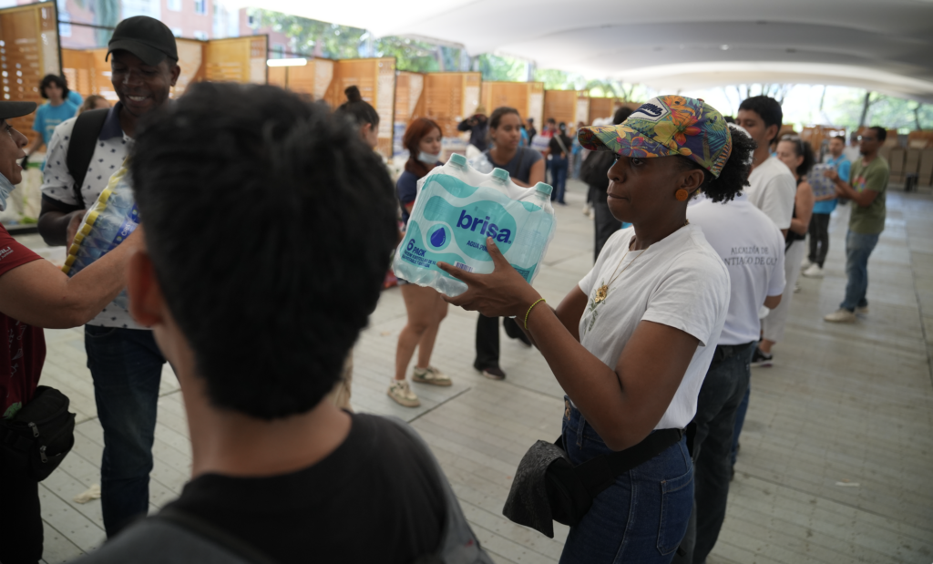 A woman passes a 6-pack of Brisa bottled water along a human chain of volunteers distributing supplies indoors