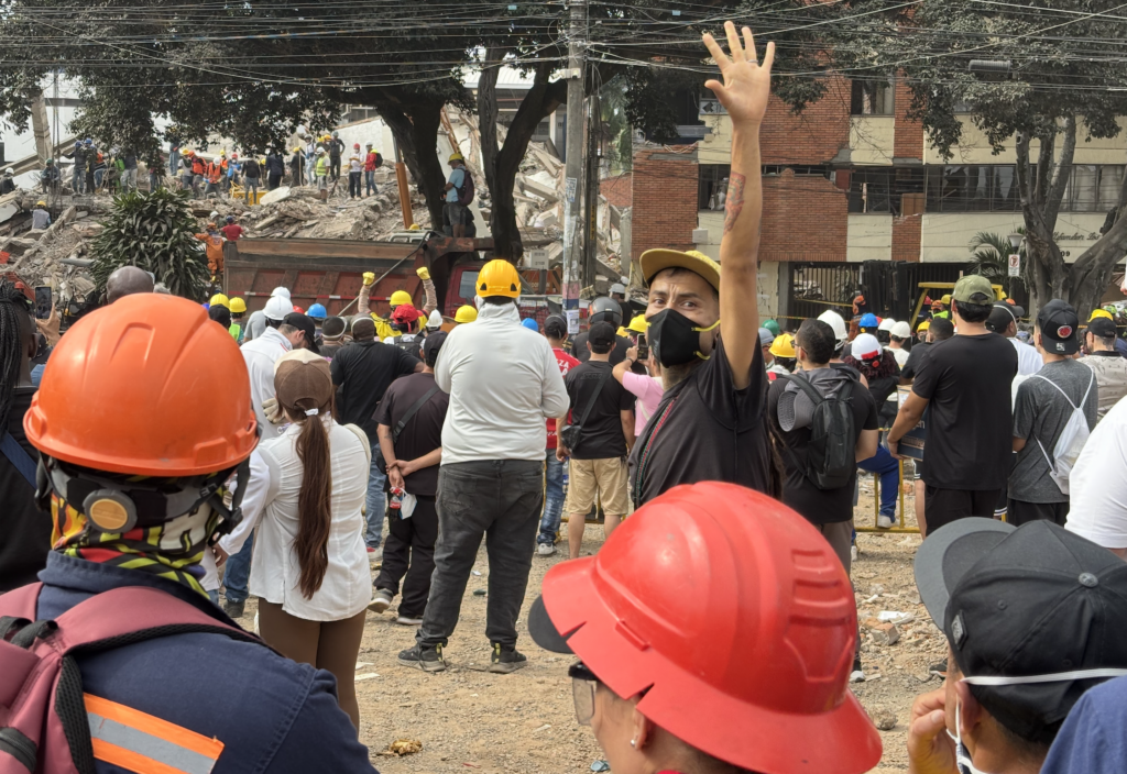 Rescue workers and bystanders gather at the site of a collapsed building, with crews searching through rubble in the background