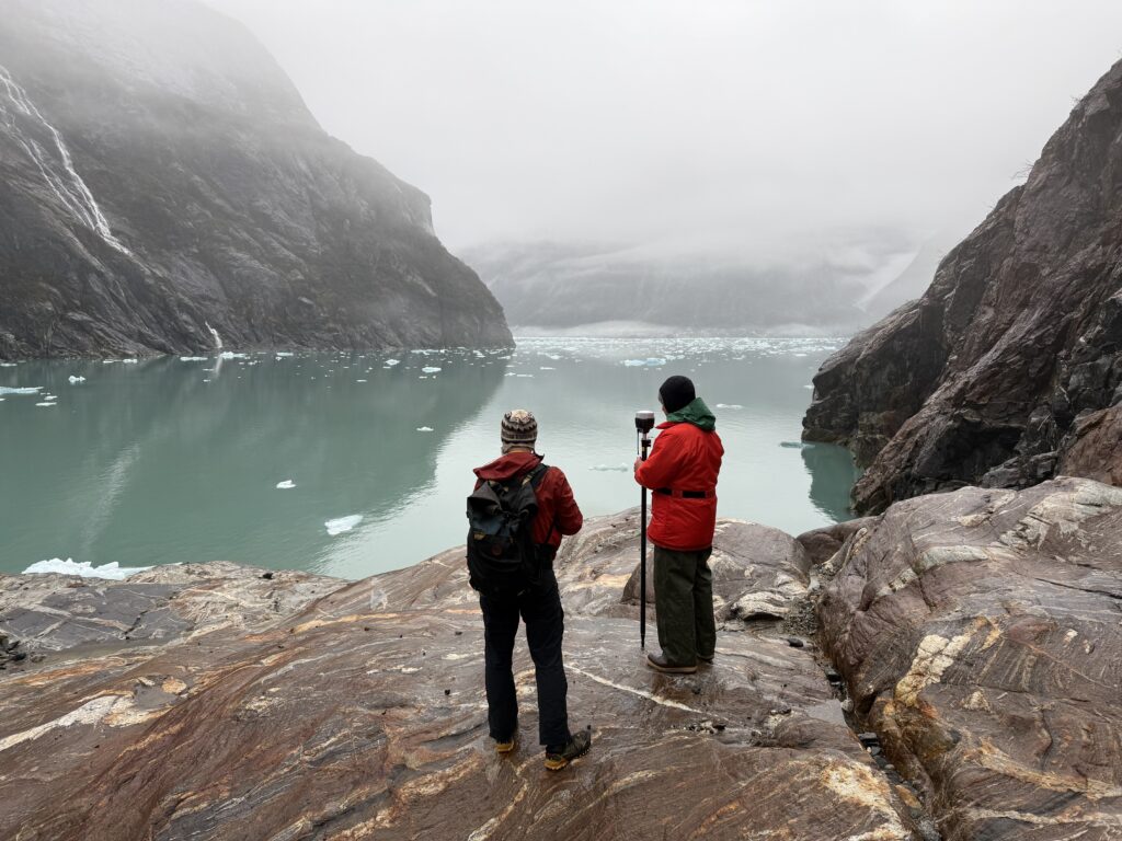 Two researchers with GPS survey equipment stand on rocks overlooking a fjord filled with floating ice, surrounded by fog-shrouded cliffs
