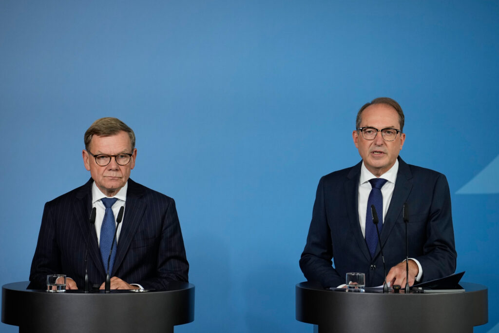 Two men in dark suits stand at separate podiums with microphones during a press conference, one speaking while the other listens