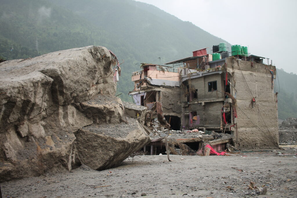 Massive boulders beside a heavily damaged multi-story building after a flood or landslide near a mountain village