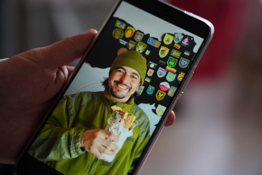 A hand holds a phone showing a smiling man in a green beanie and jacket, eating food, with military patches including Ukrainian insignia displayed behind him