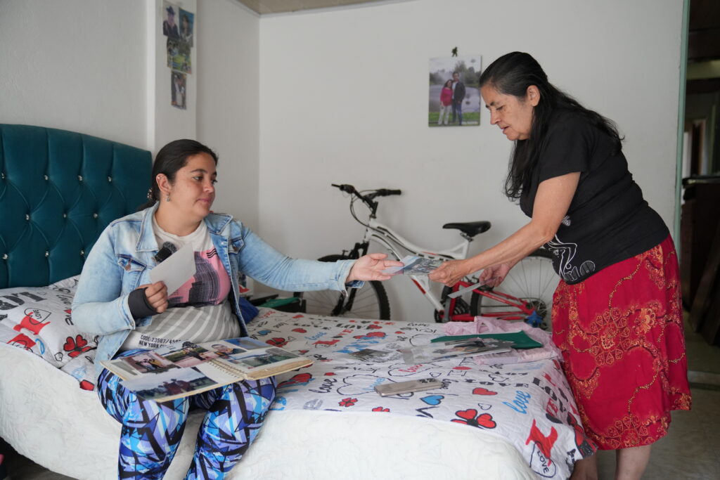 A woman hands a photograph to another seated woman looking through an album on a bed covered with family photos