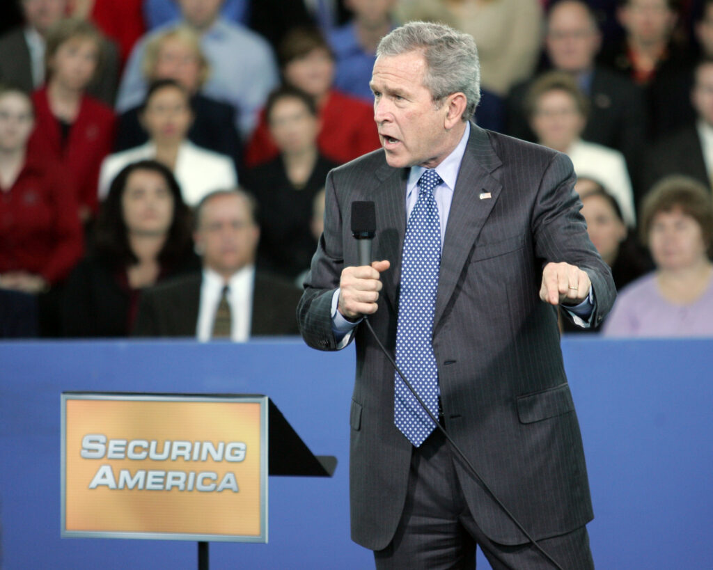 Man in suit holds microphone, gesturing while speaking to audience beside a 